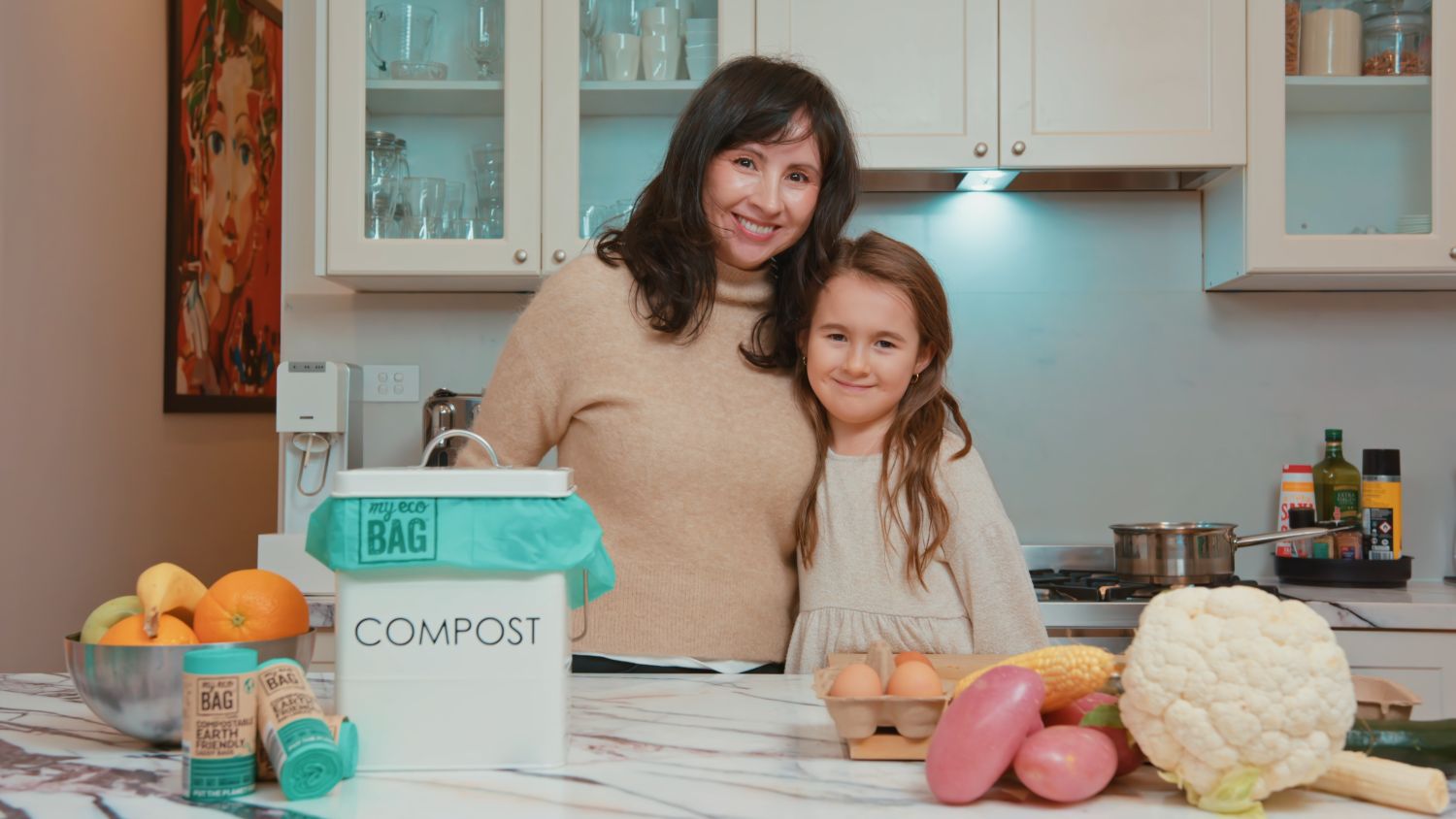 Monica Mendez stands behind a kitchen counter with her arm around a young girl. On the kitchen bench is an array of food and a compost bin with the Eco Bag product visible.